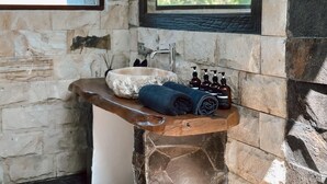 A stone bathroom with a wooden shelf holding a stone basin, towels, and toiletries. - Ocean Bay Retreat (Pecatu)