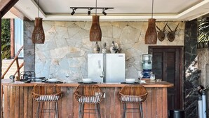 A kitchen with a stone backsplash, a refrigerator, bar stools, and hanging lights. - Ocean Bay Retreat (Pecatu)