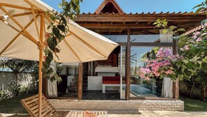 A wooden deck with a bench and an umbrella, a glass door leading to a room with a bed, and a garden with pink flowers. - Ocean Bay Retreat (Pecatu)