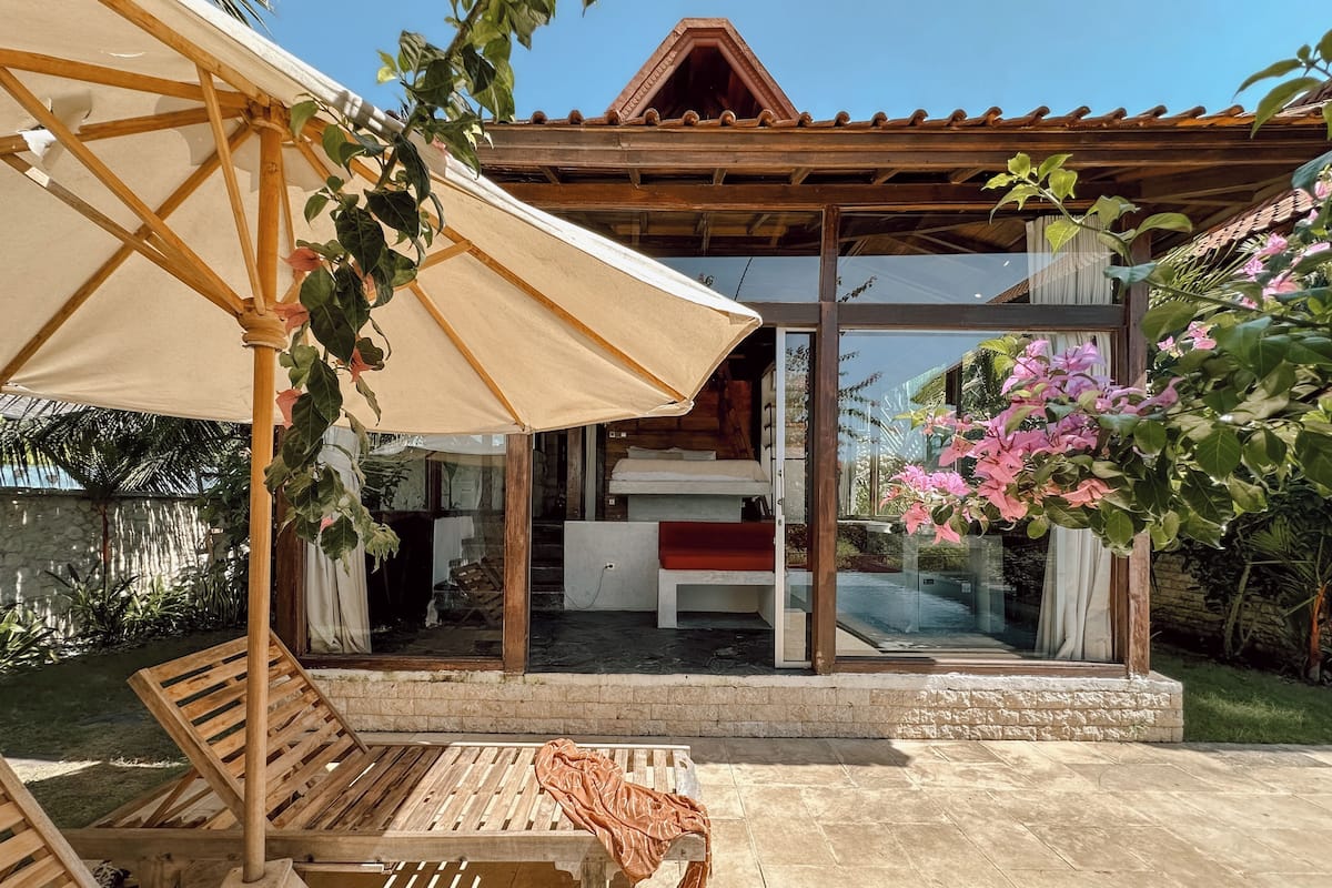 A wooden deck with a bench and an umbrella, a glass door leading to a room with a bed, and a garden with pink flowers.