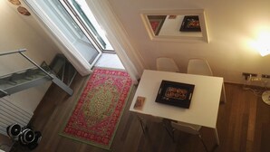 Een knus hoekje met een eettafel, stoelen, een kleedje, een plank met spullen en een raam.