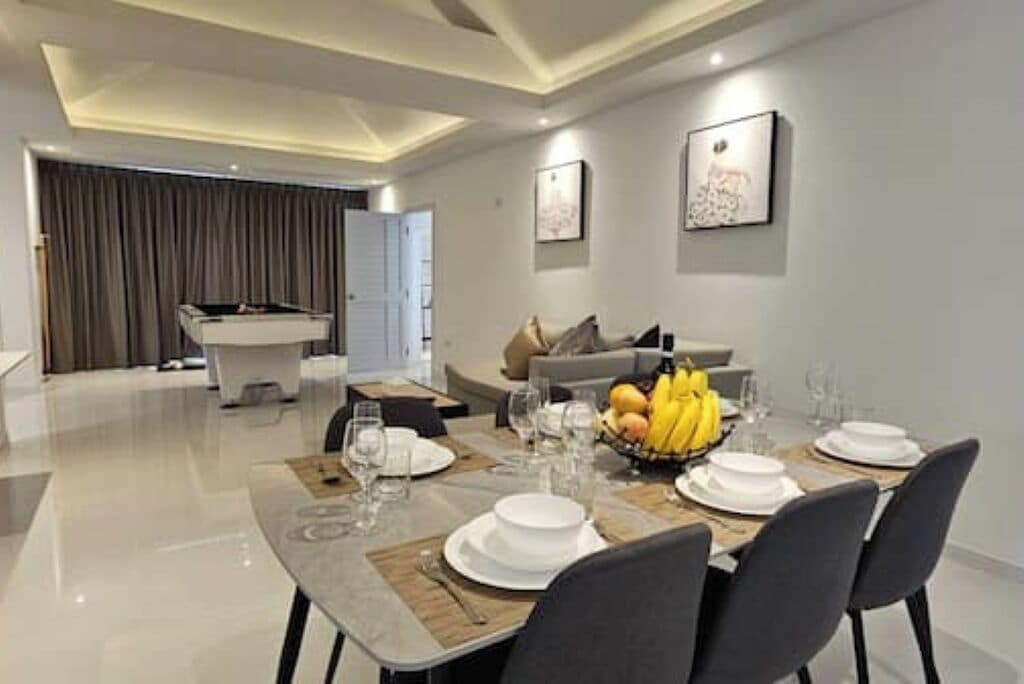 Un comedor moderno con piso pulido, una mesa de comedor preparada para cuatro personas con platos y vasos blancos, un frutero y un sofá al fondo.