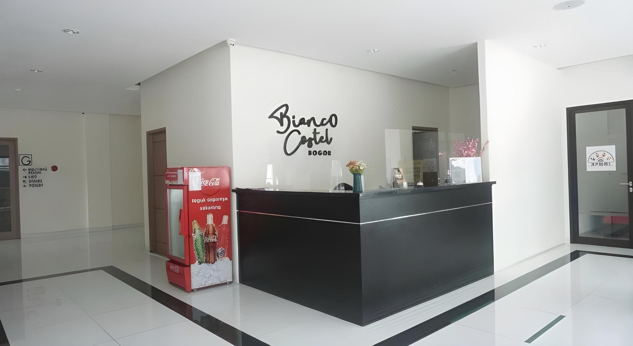 Una recepción con un refrigerador de Coca-Cola, un letrero en la pared que dice 'Bianco Castel Bogota' y una puerta de vidrio a la derecha.