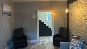 A modern living room with a staircase leading to a second floor, a black leather armchair, a grey sofa, and a textured wall. - 4 Bed Cozy Home closely located near downtown (Lansing)