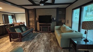 A living room with a wooden floor, a sofa, a chair, a TV, and a window with blinds. - Mountain Retreat Near Waterfalls & Hiking Trails – Your Clarkesville Getaway (Clarkesville)