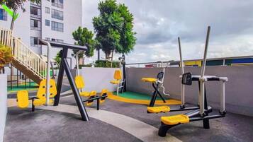 Attrezzatura per esercizi all'aperto, tra cui una sbarra per trazioni, una panca e una pressa per le gambe, in un contesto urbano con edifici e alberi sullo sfondo.