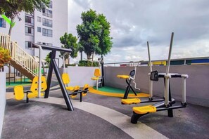 Outdoor exercise equipment, including a pull-up bar, a bench press, and a leg press, in an urban setting with buildings and trees in the background. - Sky Tree by RR JBcity (Johor Bahru)