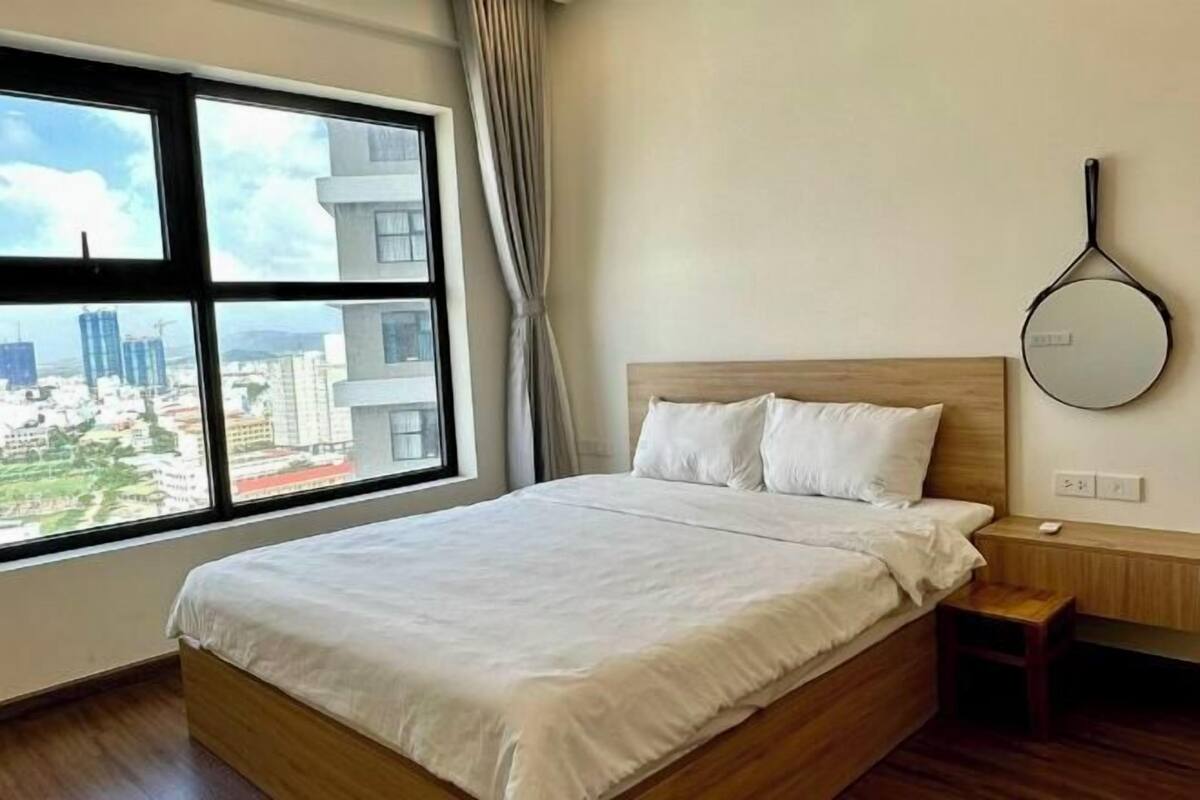 A hotel room with a large window, a bed with white bedding, a wooden headboard, a bedside table, and a wall-mounted mirror.