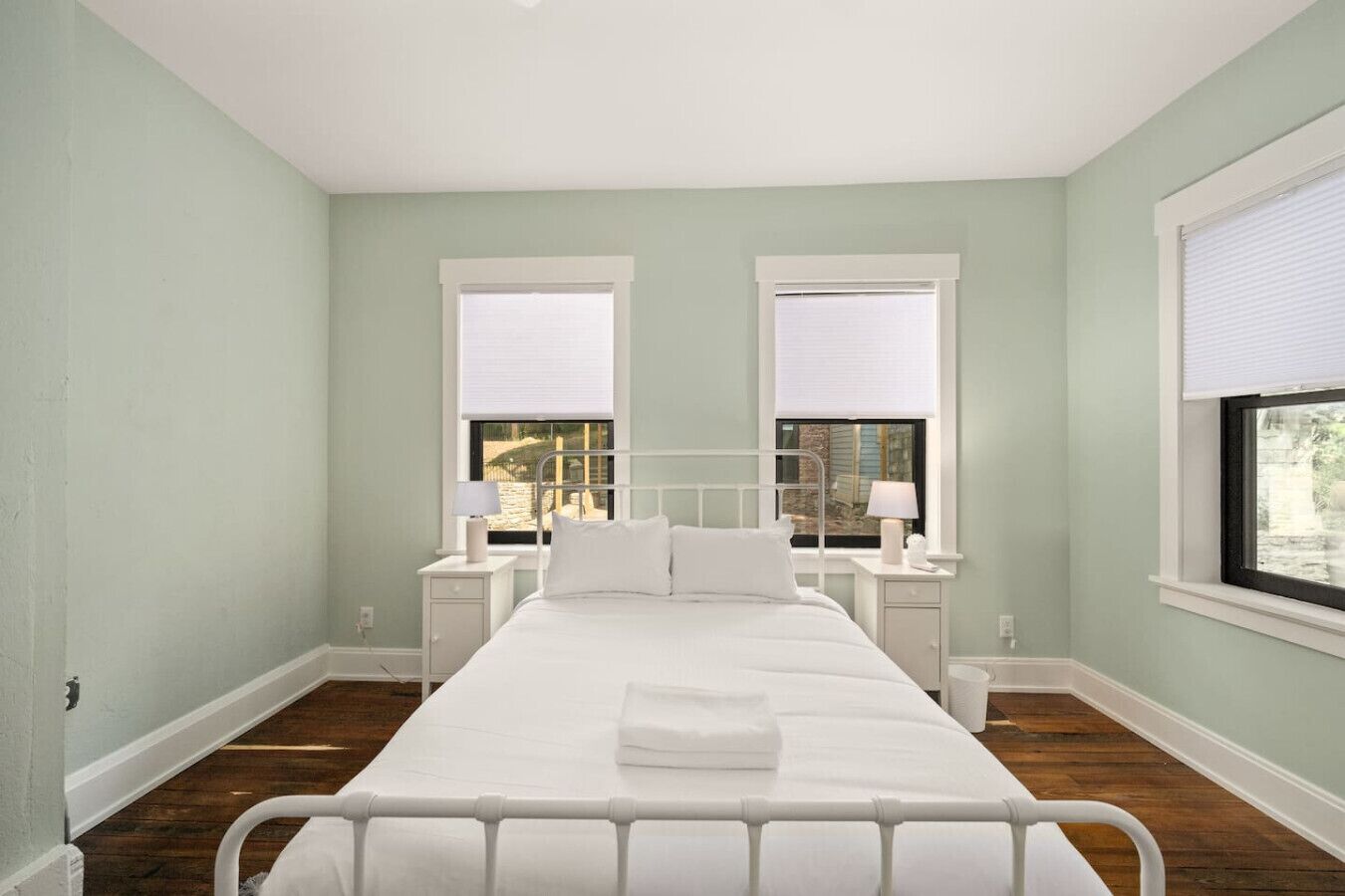 Une chambre avec un lit simple, deux tables de chevet et deux fenêtres équipées de stores.