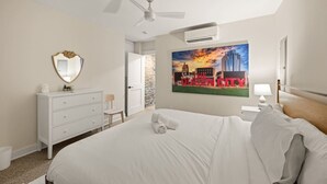 Ett hotellrum med en säng, en byrå, en spegel, en takfläkt, en luftkonditioneringsanläggning och en målning av stadsbilden på väggen.