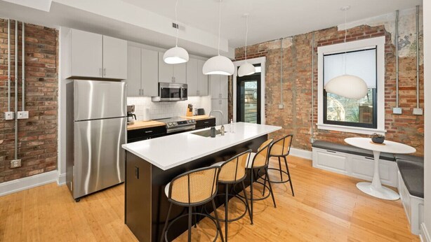 A modern kitchen with a large island, stainless steel appliances, and a brick wall. - Lux Hidden Gem, 2bd w/ Patio Steps from All OTR (Cincinnati)