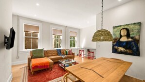A spacious living room with a large wooden dining table, a leather sofa, and a red rug. - Lux Hidden Gem, 2bd w/ Patio Steps from All OTR (Cincinnati)
