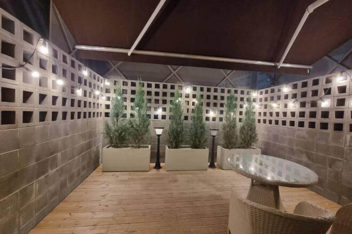 An outdoor patio area with a tiled floor, a glass table, wicker chairs, potted plants, and a lattice wall with lights.