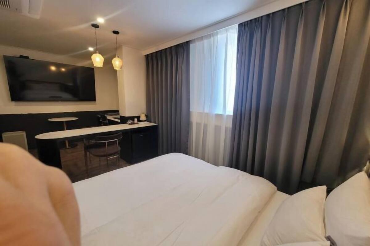 A hotel room with a bed, a desk with a chair, a TV mounted on the wall, and a window with curtains.