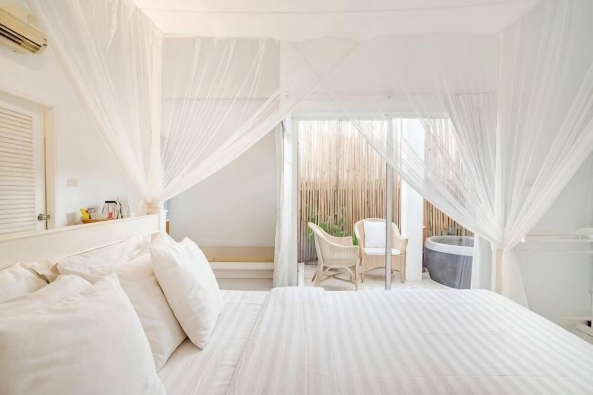Quarto com cama, cadeira, mesa pequena e janela com cortinas.