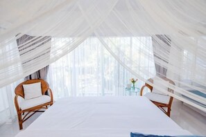 Una habitación con cama, dos sillas, una mesita con flores y cortinas traslúcidas.