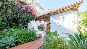 Un sendero en un jardín que conduce a un edificio con una pérgola, rodeado de exuberante follaje y plantas en flor.