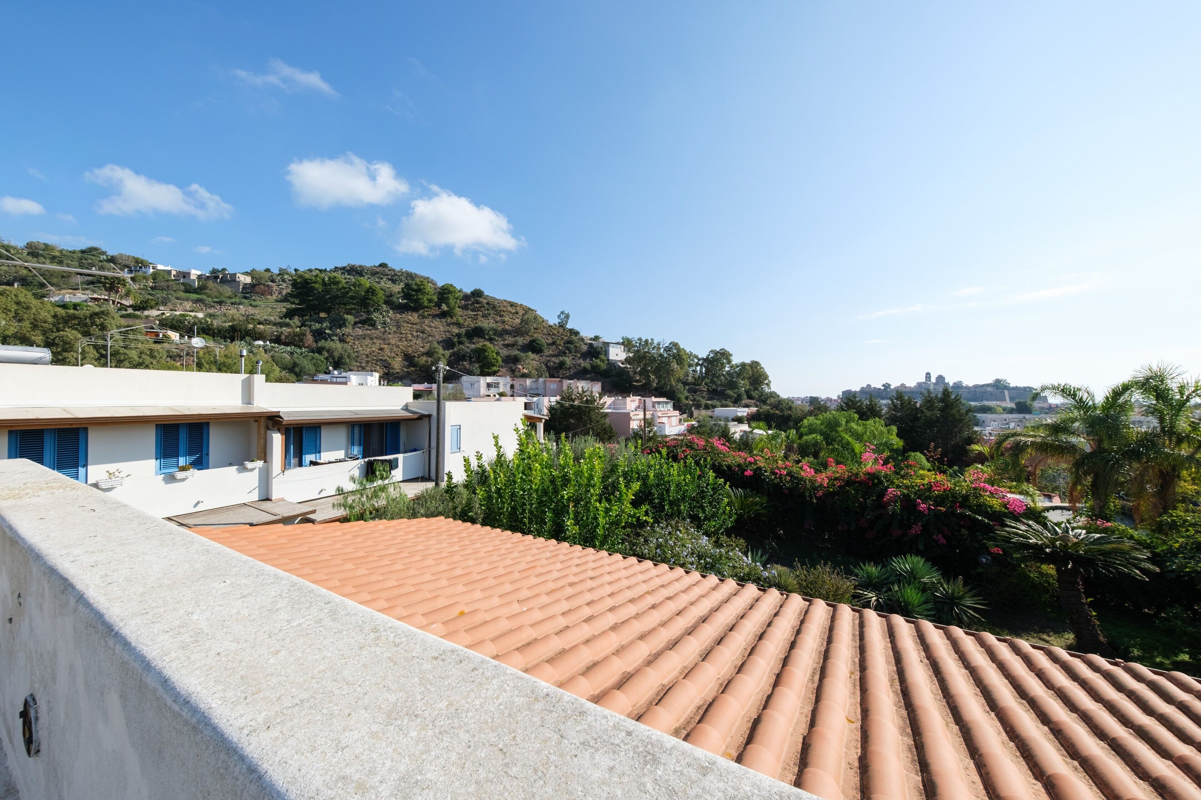 A residential area with terracotta-tiled roofs, white buildings with blue shutters, and lush greenery on a hillside.
