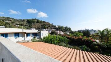 A residential area with terracotta-tiled roofs, white buildings with blue shutters, and lush greenery on a hillside.