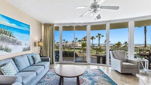 Ein geräumiges Wohnzimmer mit großem Fenster, das auf einen Strand und Palmen blickt, inklusive Deckenventilator und einem gemusterten Teppich.