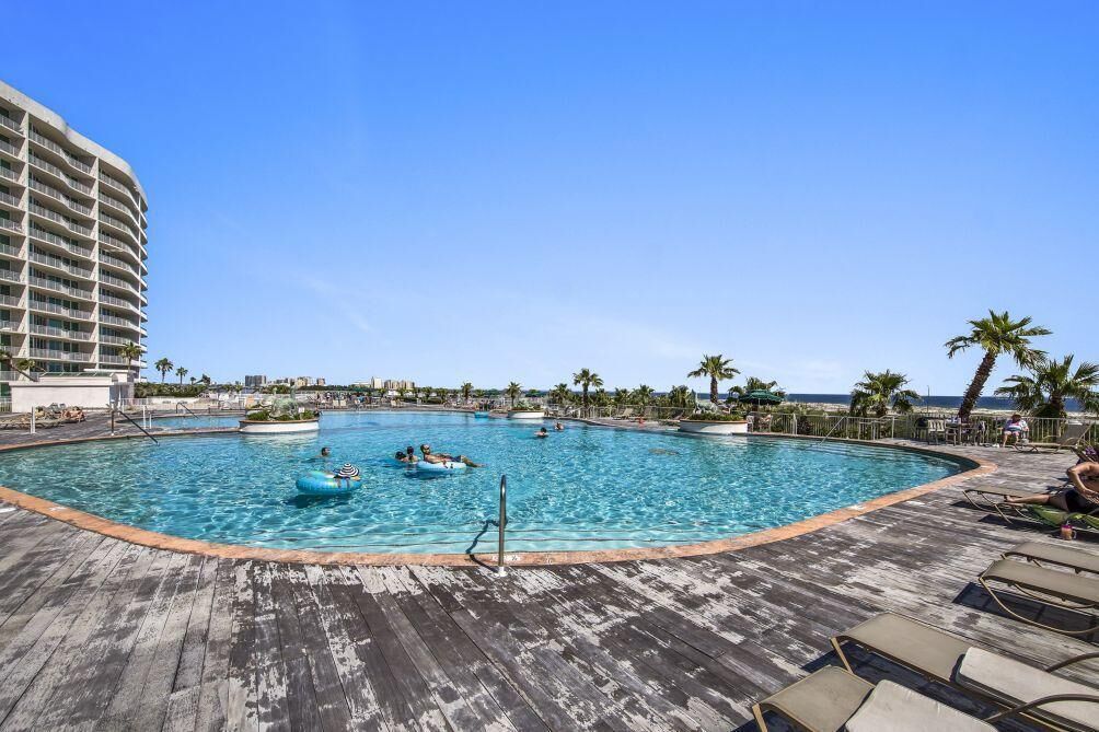A large outdoor swimming pool with lounge chairs and palm trees, surrounded by a wooden deck and a hotel building.