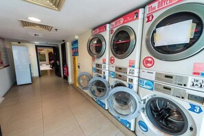A laundry facility with multiple washing machines and dryers, a tiled floor, and a visible door leading to another room. - Sunway Resort Suites by GL Staycation (Subang Jaya)