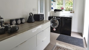 A laundry area with a washing machine and dryer, a wooden countertop with kitchenware, and a window with a view of greenery. - Roselyn House (Par)