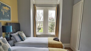 A hotel room with two beds, a nightstand with a lamp, and a window with curtains. - Roselyn House (Par)