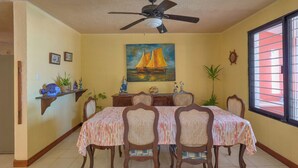 A dining room with a table set for four, a ceiling fan, a painting of a sailboat, and a window with blinds. - Casa El Cocal  -  Yucatan Home Rentals (Chicxulub)