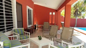 A covered outdoor seating area with white furniture and striped cushions, a small table with a plant, and a pool in the background. - Casa El Cocal  -  Yucatan Home Rentals (Chicxulub)