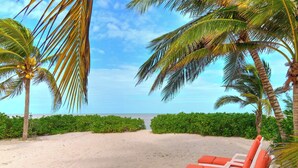 A beach with white lounge chairs and orange cushions, palm trees, and a clear view of the ocean. - Casa El Cocal  -  Yucatan Home Rentals (Chicxulub)