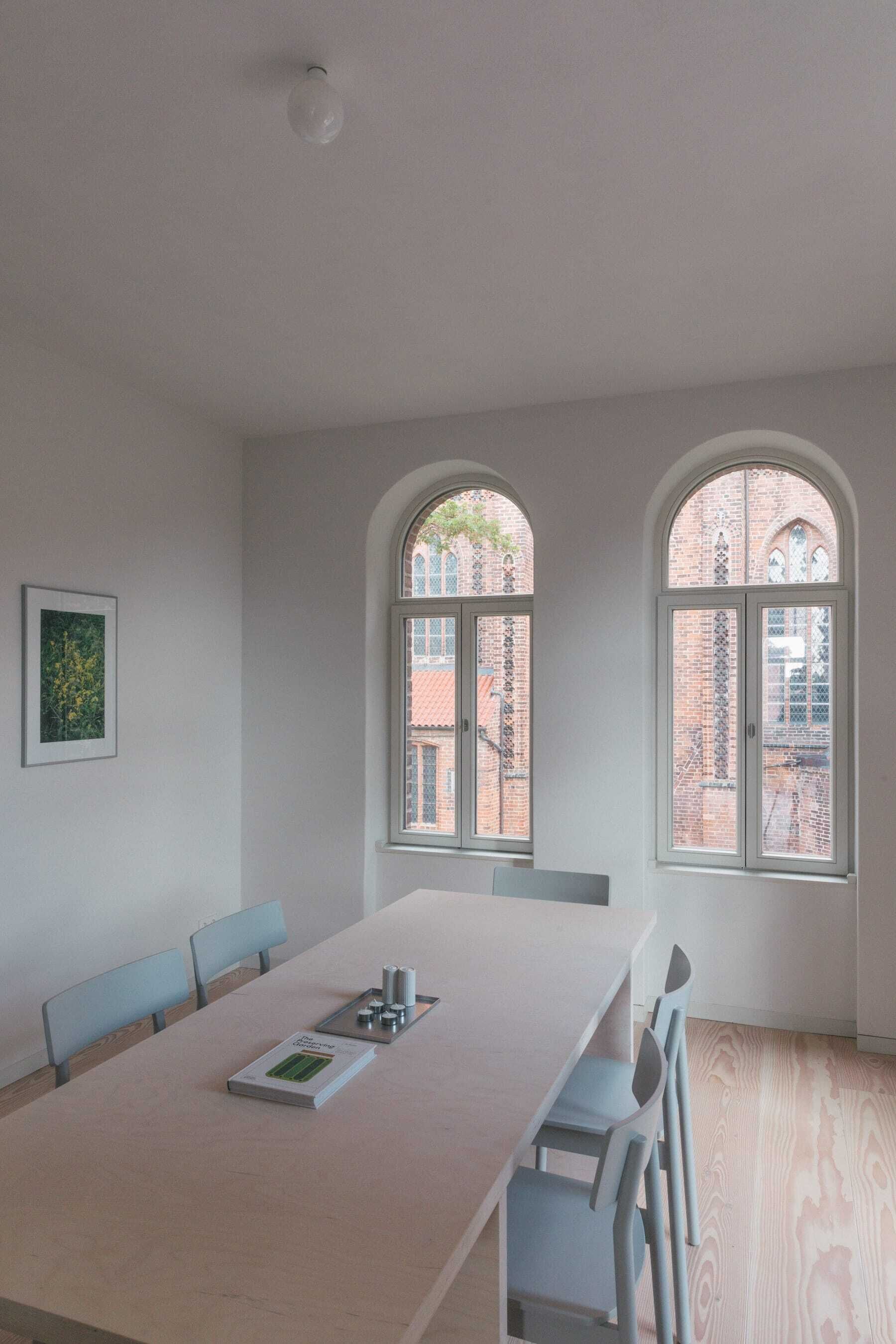 A modern dining room with a long table, chairs, and two arched windows with a view of a brick building.