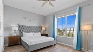 Un dormitorio con una cama grande, un ventilador de techo, una mesita de noche con lámpara, una ventana con cortinas y vistas al exterior.
