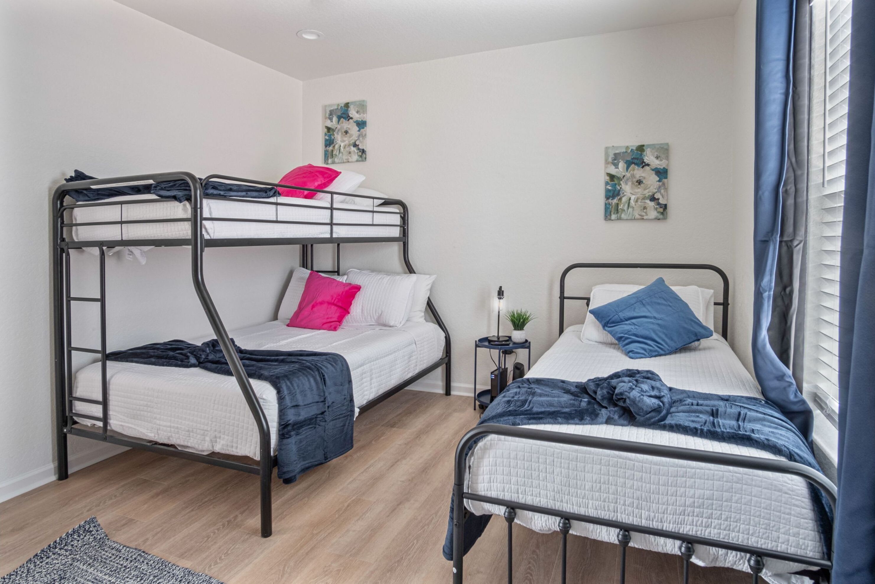 A dormitory room with two bunk beds, a small bedside table with a lamp, and floral artwork on the walls.