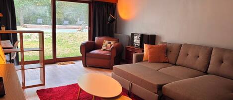 A modern living room with a sofa, a coffee table, a TV, and a large window offering a view of the outdoors.
