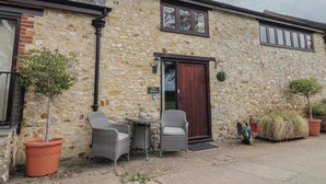 A stone building with a dark wooden door, a small table with chairs, and potted plants. - The Dairy (Axminster)