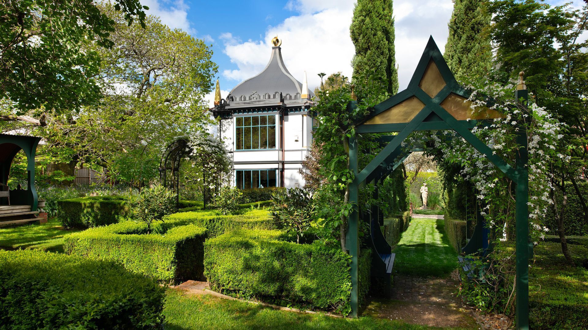 A garden with a gazebo, a white building with a pointed roof, and neatly trimmed hedges.