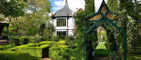 A garden with a gazebo, a white building with a pointed roof, and neatly trimmed hedges.