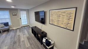 Uma sala de estar com uma TV de tela plana, um cartaz de um navio de guerra azul marinho, um sofá e piso de madeira.
