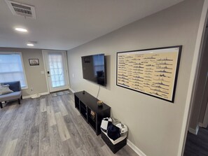 A living room with a flat-screen TV, a navy ship poster, a sofa, and a wooden floor. - Charming 3-bedroom Barrancas Bungalow in Pensacola with AC (Pensacola)