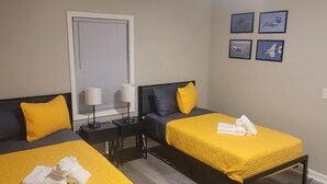 Quarto de hotel com duas camas, cada uma com colcha e travesseiros amarelos, uma mesa de cabeceira com abajur e uma janela com persianas.