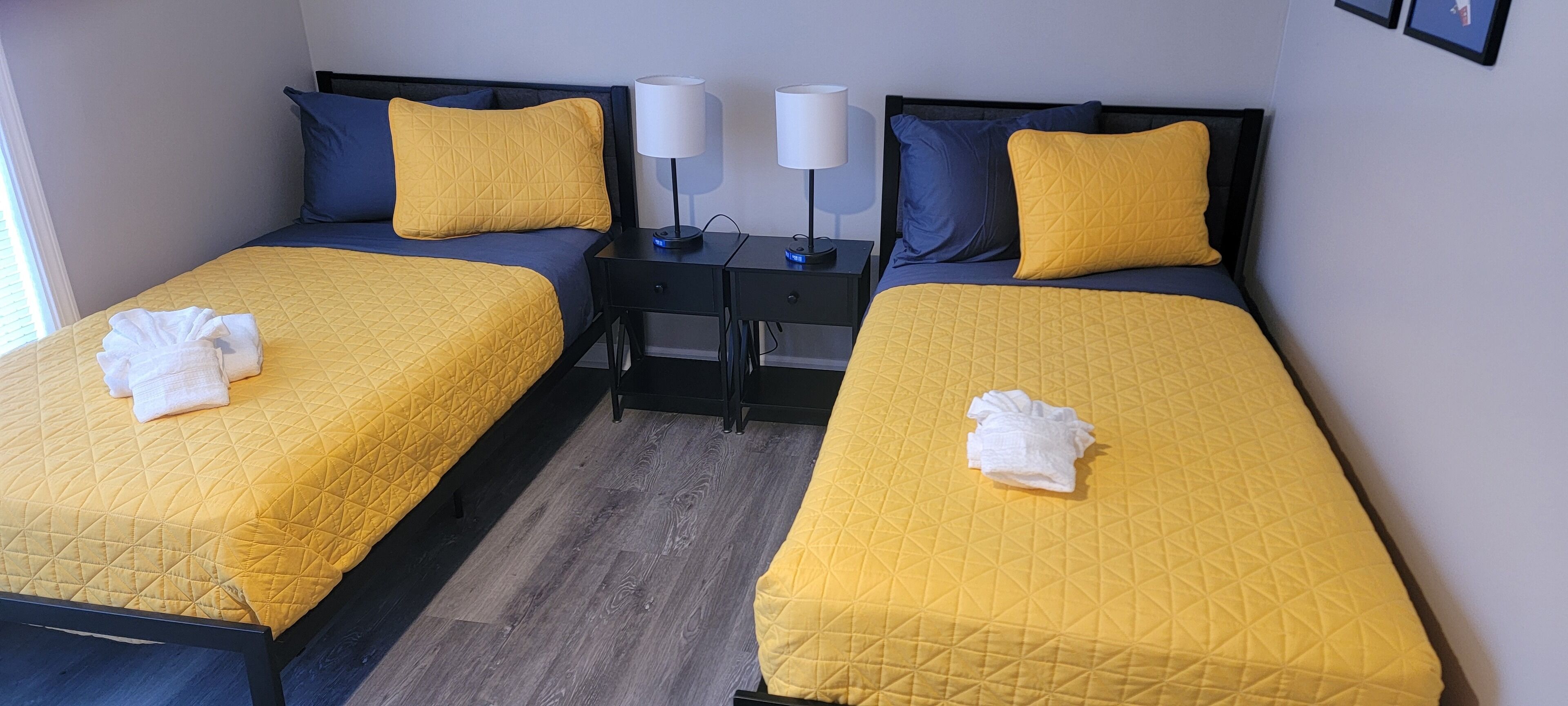 Ein Hotelzimmer mit zwei Betten, jedes mit einer gelb gepolsterten Bettdecke und einem auf dem Bett gefalteten weißen Handtuch. Es gibt zwei Nachttische mit Lampen und eine kleine Schublade.