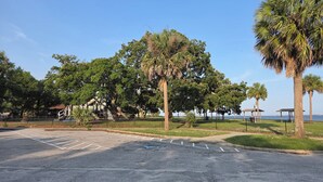 Um estacionamento com vagas demarcadas, uma área ajardinada com árvores e vista para o mar.