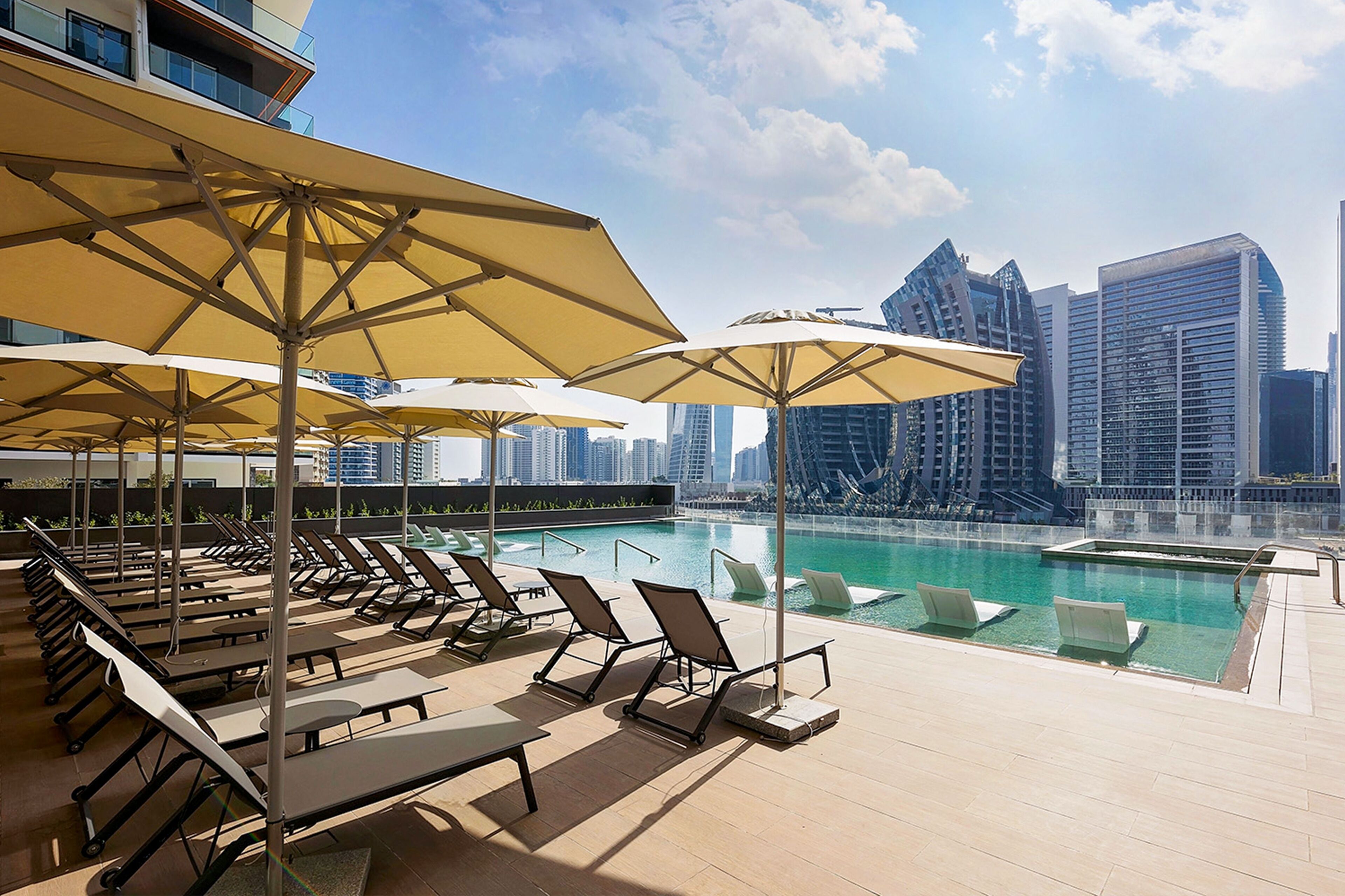 A rooftop pool area with sun loungers and umbrellas, overlooking a city skyline.
