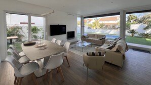 A modern living room with a dining area, large windows, a flat-screen TV, and a sofa. - Villa Das Hortensias (Aroeira)