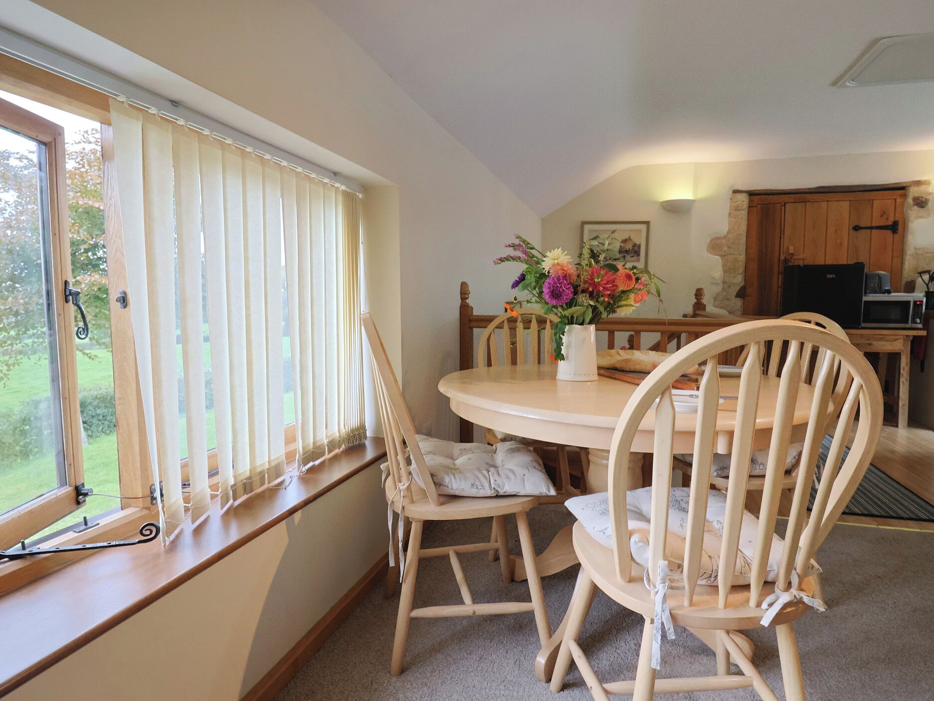 Comedor con una mesa redonda, sillas de madera, un florero con flores y una ventana con cortinas.