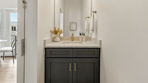 Een moderne badkamer met een witte wasbak, donkere kastjes en een grote spiegel. Boven de spiegel hangen drie lampen en op het aanrecht staat een klein plantje.