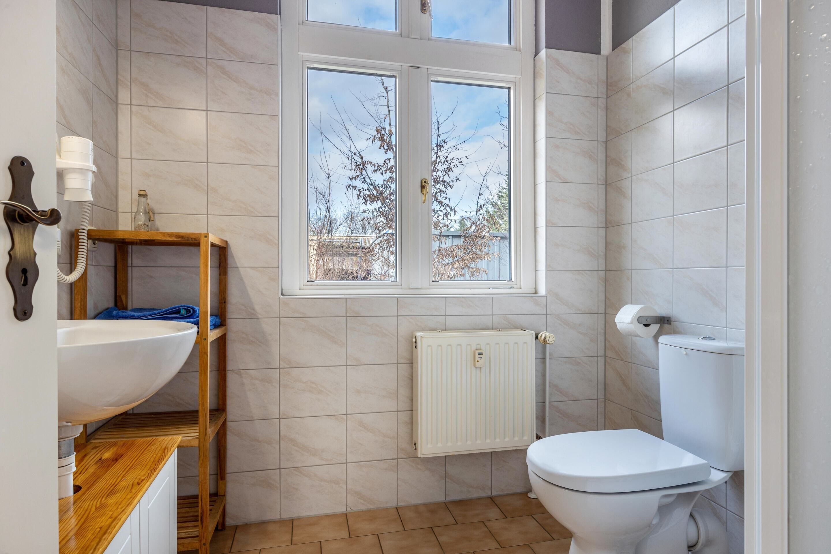 A bathroom with a white toilet, a wooden shelf, a white sink, and a window with a view of trees.