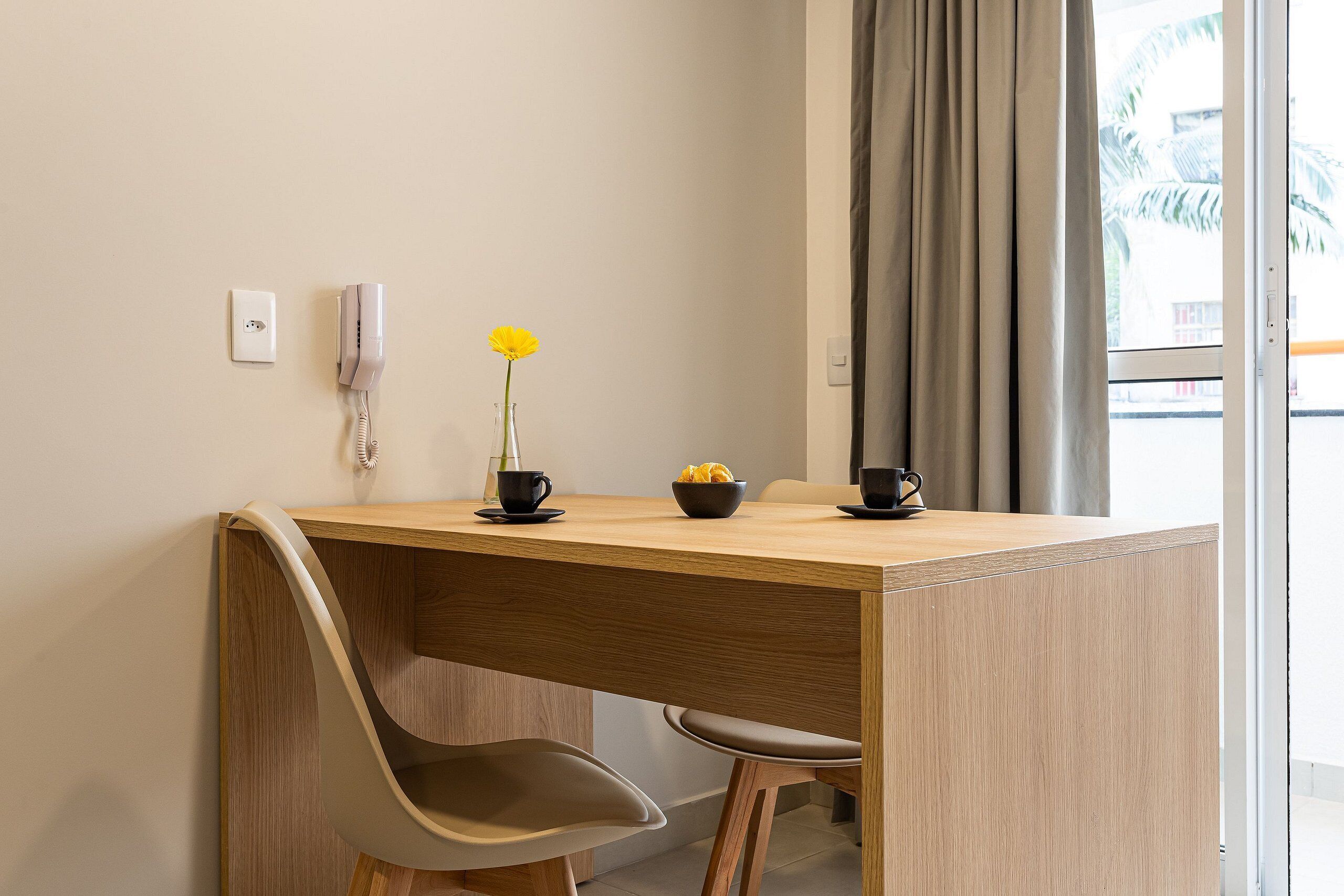 A small, modern room with a wooden desk, two chairs, a vase with a yellow flower, and a bowl of fruit.