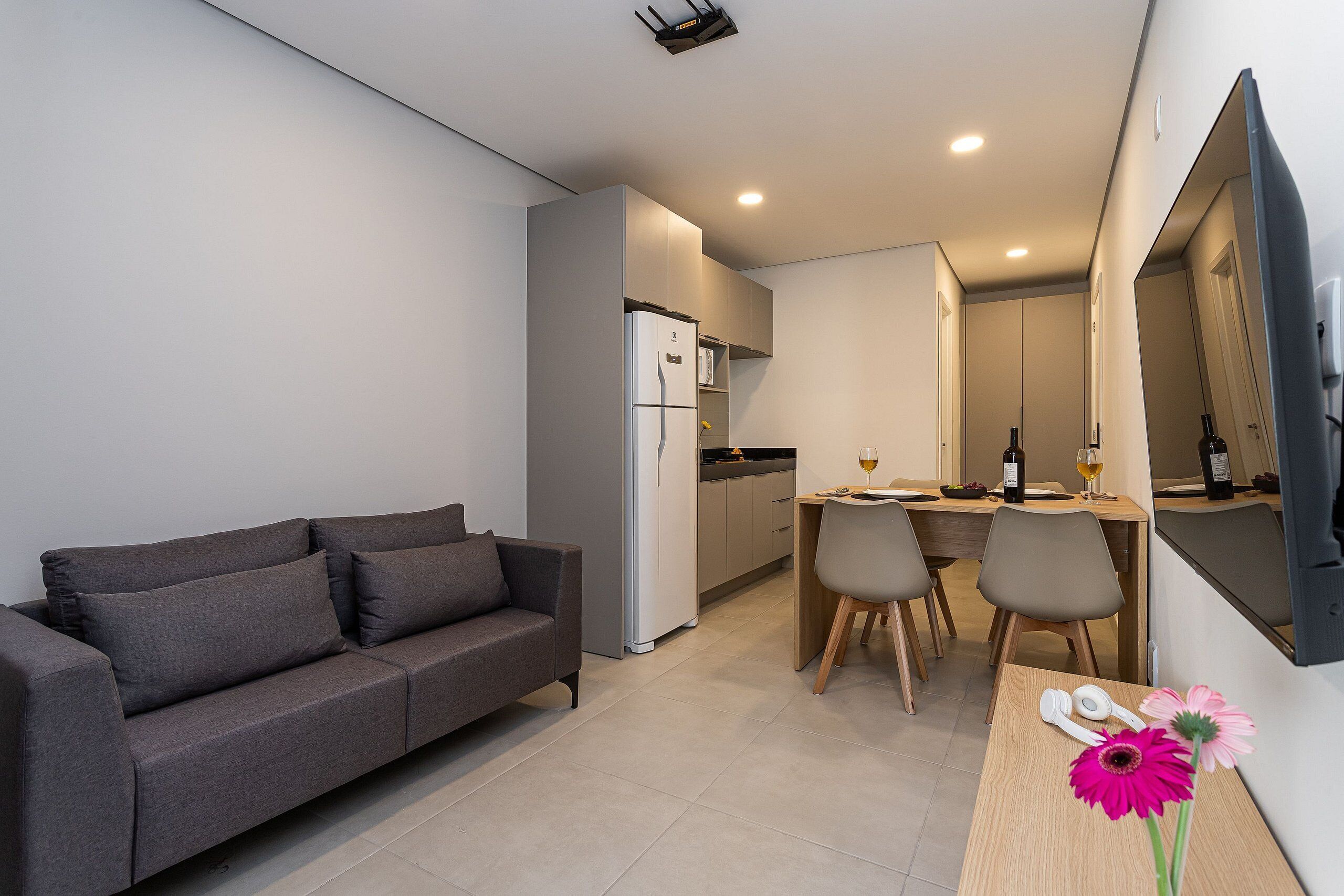 Una sala de estar moderna con un sofá gris, un comedor con dos sillas, una cocina con refrigerador y un televisor montado en la pared.